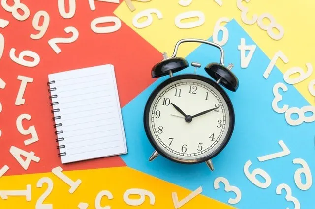 Relógio despertador rodeado de papel multicolor e um caderno em branco, com números brancos dispersos 