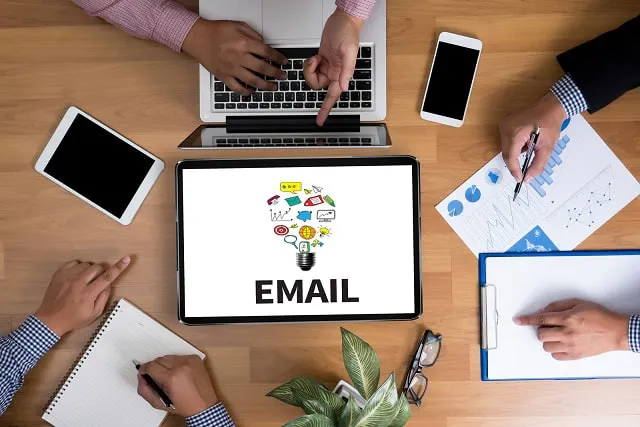 Ein Team arbeitet an einem Tisch mit einem Tablet, auf dem eine E-Mail-Grafik mit Symbolen in Form einer Glühbirne abgebildet ist.